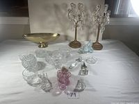 Overview of entire lot showing the mixed items spread on a white tablecloth under light, including candle holders, crystal dishes, wine stoppers, and a gold-toned bowl.