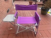 Front and side angled view of purple folding outdoor chair with attached translucent tray table and padded armrest on red wooden deck.