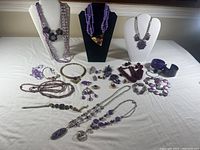 Wide view of various purple costume jewelry displayed on stands and flat on table including necklaces, bracelets, earrings, and pins.
