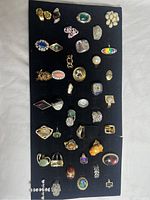 Photo showing 36 assorted costume jewellery rings laid out on black felt board, including various shapes, colors, and metal tones.