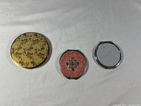 Set of three vintage compacts: gold-tone makeup compact with decorative lid, small silver-tone mirror compact, and round silver-tone mirror compact with textured back.