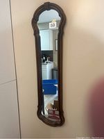 Full view of tall narrow mirror with ornate wooden frame hanging on wall, showing surrounding room reflection.