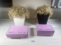 Two blonde curly wigs on mannequin heads with their respective Paula Young purple boxes placed in front of them.