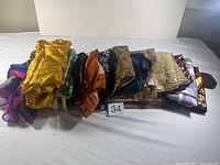 Photo showing thirteen scarves arranged side-by-side, displaying various colors and patterns including gold, dark, multicolor, and sheer fabrics.