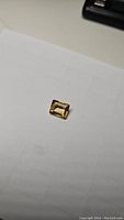 Photo of a small emerald cut deep golden citrine gemstone placed on white paper, showing its color and facet pattern.