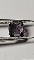 Close-up photo of a cushion-cut purple pink spinel gemstone held by tweezers, showing its faceted surface and color.