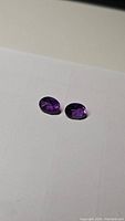 Pair of deep purple oval cut amethyst gemstones placed on a white background, showing size and color consistency between stones.
