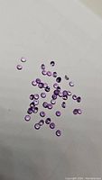 Close view of multiple small round calibrated purple amethyst gemstones scattered on a white surface showcasing their diamond cut facets and purple color.