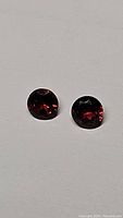 Close-up of two round faceted cherry red garnet gemstones on a neutral background, showing deep red color and diamond cut facets.