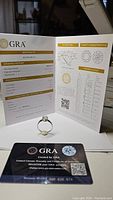 GRA certificate and authenticity card displayed behind the ring, certifying the 1 ct moissanite with VVS1 clarity and D color.