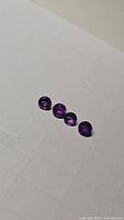 Four round, faceted deep purple amethyst gemstones laid on white surface, visible cut and clarity.