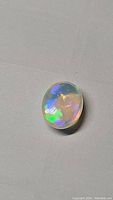 Photo of oval cabochon cut natural Ethiopian opal on a grey surface showing bright blue, green, and yellow flash colors.