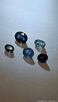 Five oval cut sapphires arranged in a row on a white surface with visible shadows and light reflections, showing varying blue and multicolored hues, showcasing size and color differences.