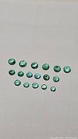 Top view of 18 natural Zambian emeralds arranged in three rows on a light background, showing varying sizes and emerald green color.