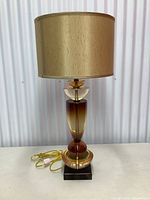 Full front view of lamp showing amber gradient glass body, gold metal accents, black square base, and drum shade