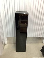 Front view of tall black plexiglass pedestal with a reflective surface, showing some scratches and scuffs.