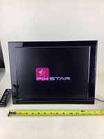 PixStar digital picture frame powered on, displaying logo, with measuring tape showing width around 16 inches.