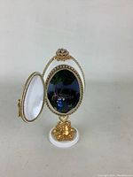 Front view of the Faberge-style egg opened showing the interior 3D scene of a couple in a garden with bench and tree, white and gold gilt with pearl trim.