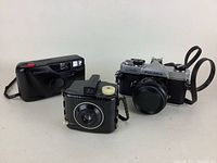 Front angle view of three cameras: black Samsung point-and-shoot, black box camera with yellow knob, Fujica ST605 film SLR camera with lens cap and strap