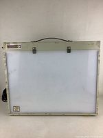 Front view of the vintage portable light box showing white illuminated panel and cream-colored metal frame with handle and clips.