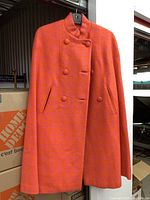 Photo showing the full cape hanging, highlighting its orange and pink houndstooth pattern, sleeveless design, and double-breasted button front.