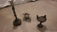 Three pewter candle holders with intricate cluster designs placed on a floor background.