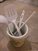 Four-piece kitchen set: ceramic crock holding three white kitchen tools including slotted spatula, solid spoon, and stirring spoon all branded Chefmate.