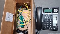 Photo showing the AT&T two-line corded speakerphone with numeric keypad, function buttons, and a digital display alongside a cardboard box containing various telephone cables in yellow, green, and black.
