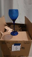 Single deep blue wine glass placed on a cardboard box, showing its tall stem and rounded bowl.
