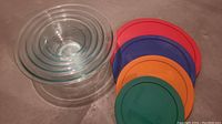 Five nested clear glass Pyrex baking bowls alongside five colorful plastic lids on a flat surface.