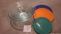 Set of nested clear glass Pyrex bowls with four colorful plastic lids on the side, viewed from above showing nested bowls and lids.