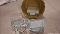 Overview of all items including the round metal baking pan with lid, two square glass Pyrex dishes with blue lids, and clear glass measuring cup.