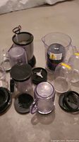 Magic Bullet blender base unit along with multiple clear plastic cups, screw-on blades, lids and a measuring jar on a beige surface.