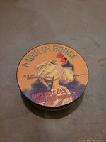 Round box with vintage Moulin Rouge artwork depicting a can-can dancer, labeled 'Set of Four Salad Plates'. Orange background with Moulin Rouge in large letters.