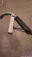 Two power strips, one white and one black, lying on concrete floor showing multiple outlets and attached power cords