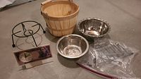 Various kitchen and household items arranged on floor including wooden basket, metal plant stand, three metal bowls, small wire rack in plastic bag, and decorative clock.