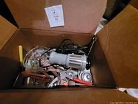 Box filled with assorted kitchen utensils including metal and plastic handled items: rotary grater, scissors, cutters, whisks, strainer, forks, spoons, and spatulas.