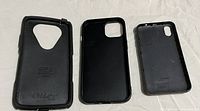 Photo showing three black iPhone cases, including one OtterBox case and two other black protective cases, placed side by side on a light-colored fabric surface.