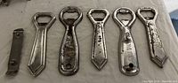 Photo of six vintage metal bottle openers laid out in a row on a white cloth background showing different shapes and wear levels.