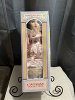 Porcelain doll in pink satin dress, face and hair visible through window of original Cathay Collection box.
