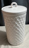Image of a tall white ceramic canister with a textured diamond surface and round loop handle lid.