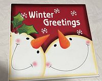 Photo shows a square Christmas wall plaque with two smiling snowmen, orange carrot noses, holly leaves, berries, and 'Winter Greetings' text on a red background with snowflakes.