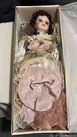 Porcelain doll dressed in pink and cream lace dress inside white box, showing doll's face, hair, and dress details