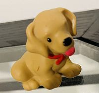 Photo of a small yellow dog figurine from Little People, placed on a reflective surface. The dog has black eyes, a black nose, a pink tongue sticking out, and a red collar around its neck.