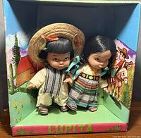 Two Lupita dolls in traditional attire displayed inside original box with illustrated Mexican countryside background.