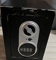 Front view of mini black safe showing electronic keypad with circular silver trim and manual keyhole below it. Safe door and edges show wear and chipped paint.