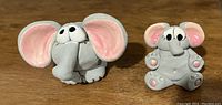 Photo of two small stone elephant figurines with pink ears and black eyes, placed on a wooden surface.