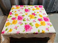 Photo of a square tablecloth spread out, showing a floral pattern with pink and yellow flowers and green leaves on a white background.