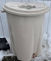 Beige cylindrical rain barrel with lid, spigot near base, placed outdoors on snow. Crack at bottom visible.