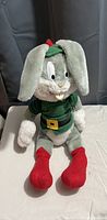Vintage Bugs Bunny plush doll sitting on white sheet in front of gray background, showing front view with character in detailed green outfit and red feet.
